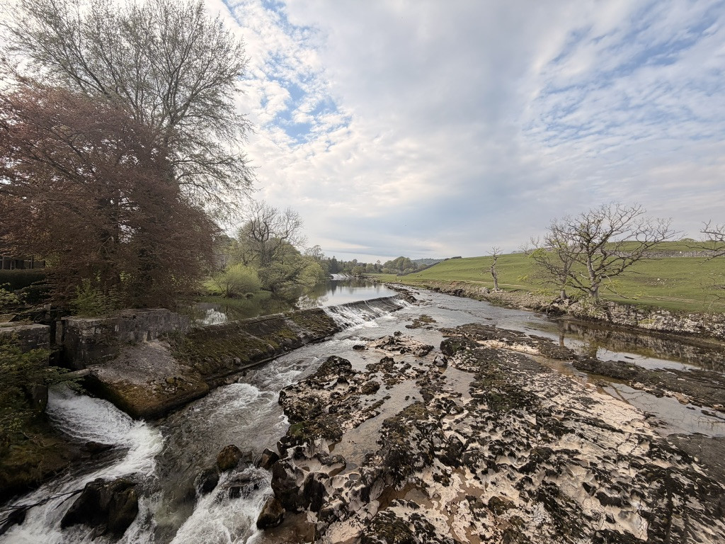 Wide view of the River Wharfe flowing over Linton Falls beneath a bright, cloud-filled sky