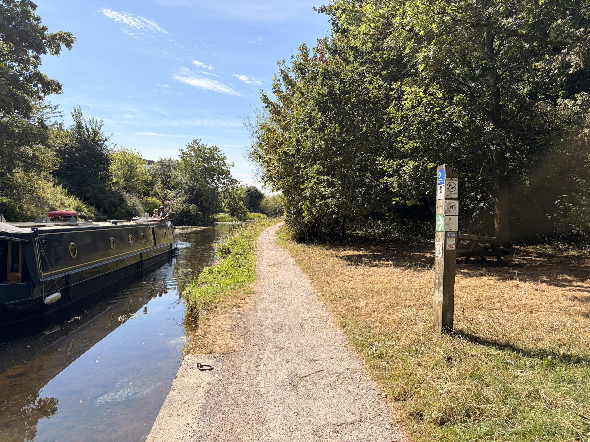 Narrowboat travelling beside a sunny tree-lined towpath on the Lancaster Canal