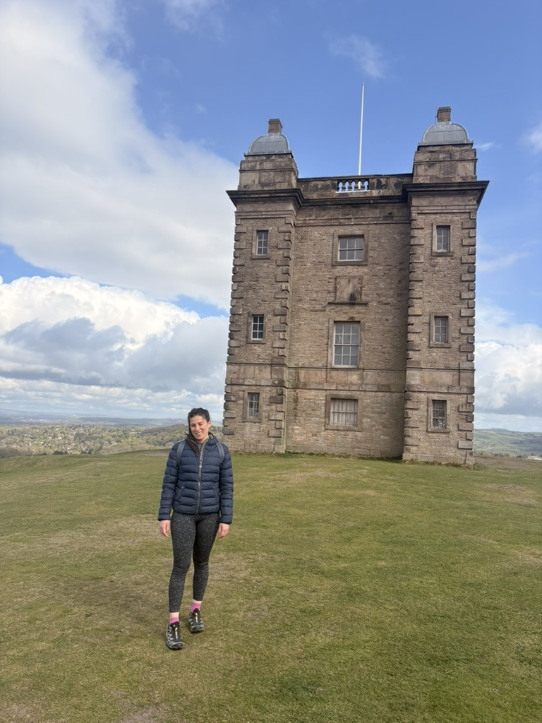 Veronica standing on the grass in front of The Cage at Lyme Park, with the stone tower rising behind her beneath a blue, cloud-filled sky.