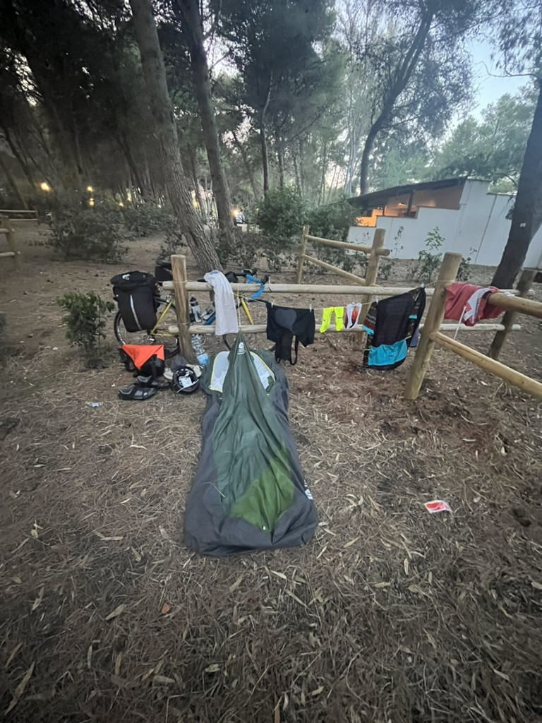 A compact one-man tent beneath the trees at Torre dell’Orso, with a loaded touring bike and cycling kit drying on a wooden fence.
