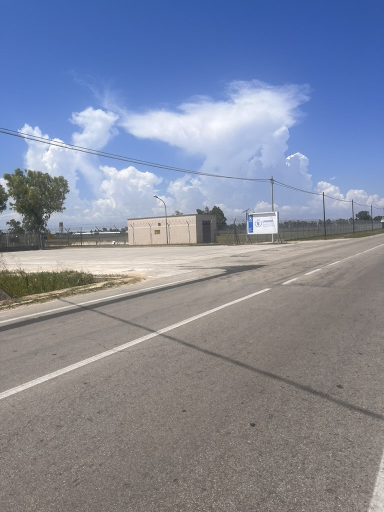 A roadside view of the fenced UN Humanitarian Response Depot in Brindisi beneath towering white clouds.