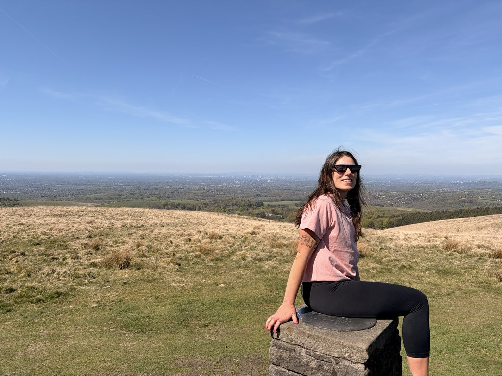 Veronica sitting on the stone trig point at the top of Lyme Park, with open moorland and expansive views over Cheshire beneath a clear blue sky.