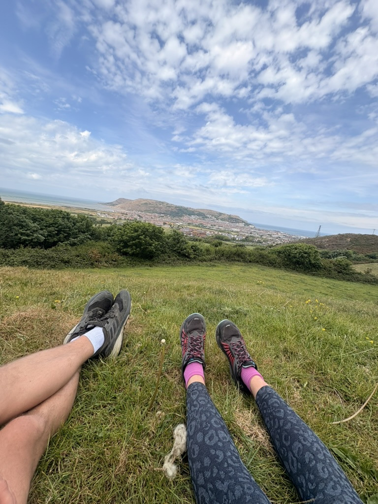 Two hikers resting on a grassy hillside with Llandudno and the sea spread out below.