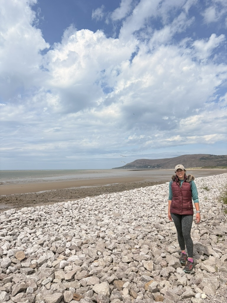 Veronica standing on a rocky beach beneath a wide, cloud-filled sky.