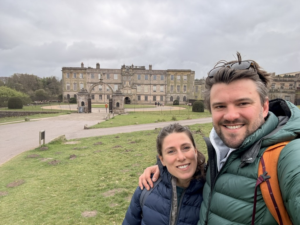 Edward and Veronica smiling for a selfie on the grass in front of Lyme Park house beneath an overcast sky.