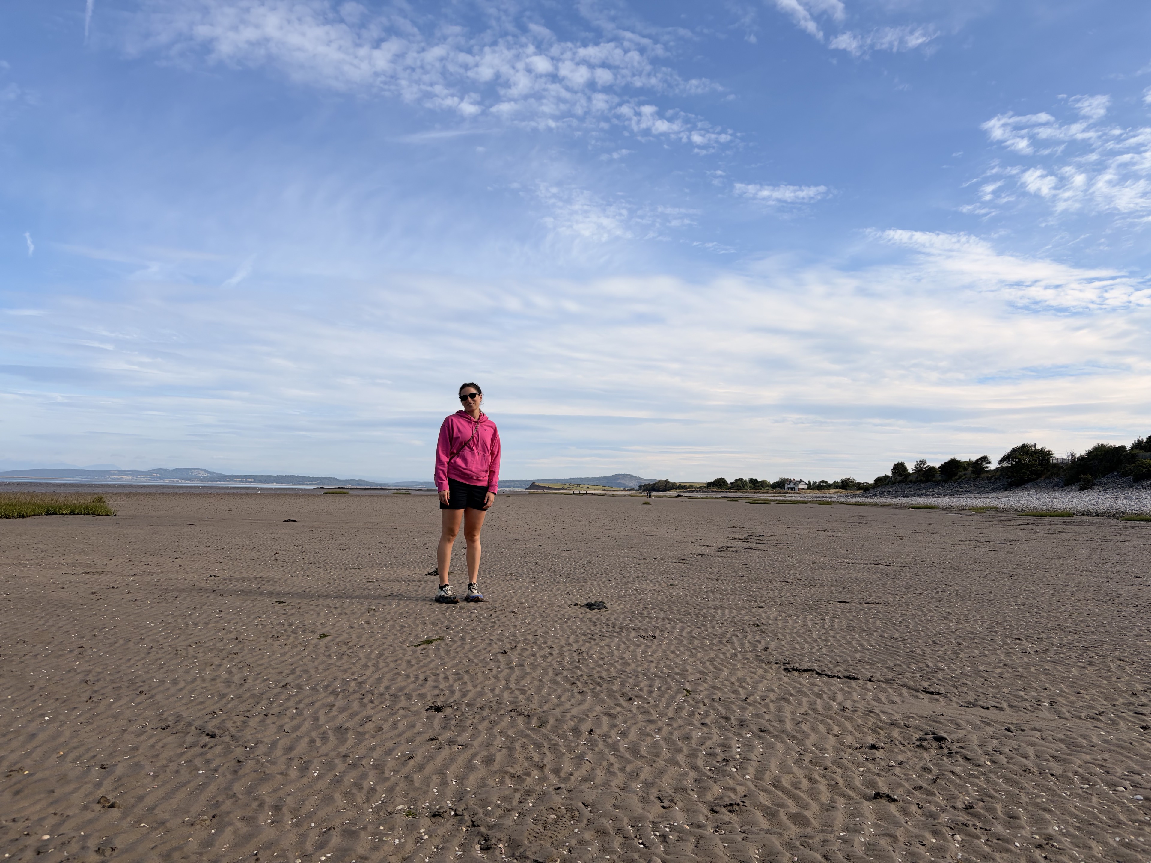 A walker in a pink jacket standing on the vast exposed sands of Morecambe Bay beneath a wide blue sky.