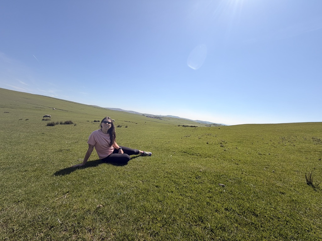 Veronica sitting in a broad, sunlit grass field above Lyme Park, with sheep and distant Peak District hills under a clear blue sky.